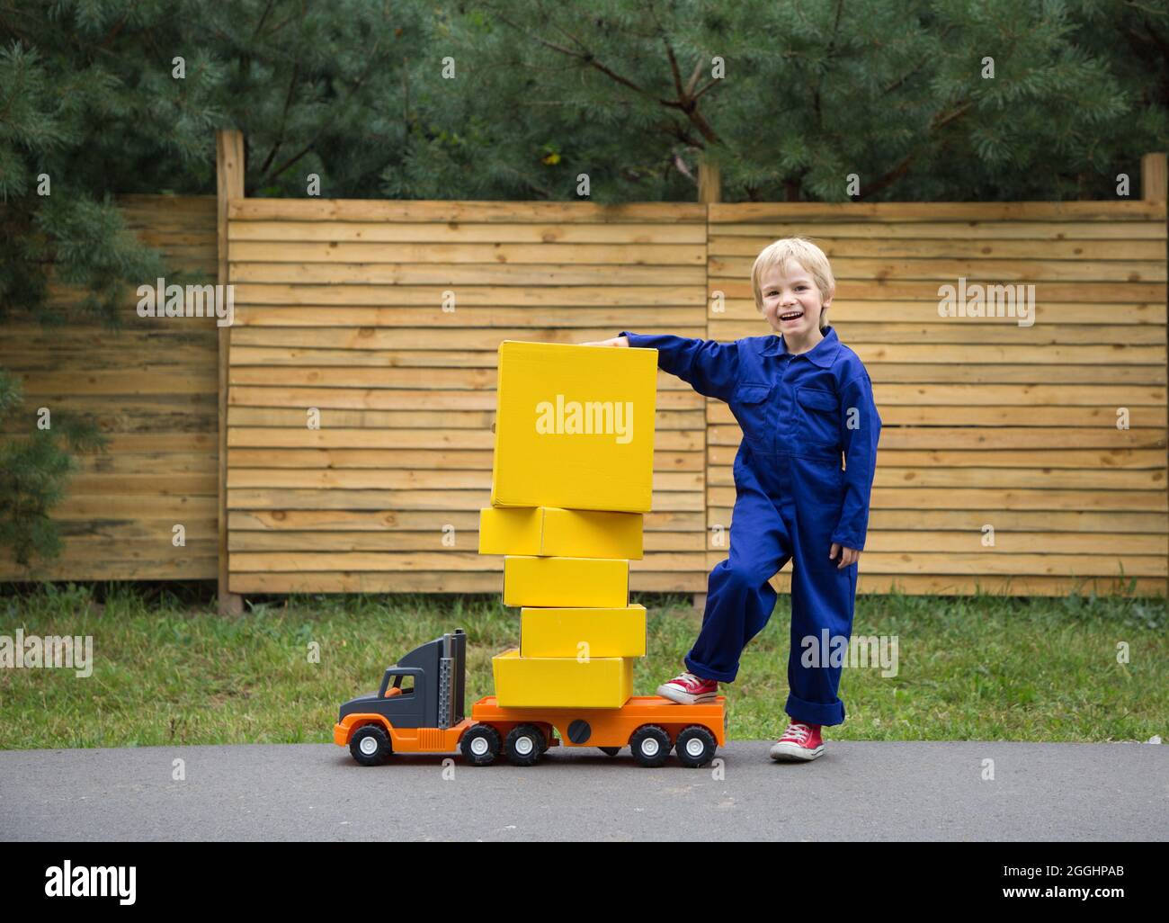 un niño de 5 años de edad en un mono azul se encuentra cerca de un gran  coche de juguete - un camión, cargado con cajas de cartón amarillo. Poco  mensajero en