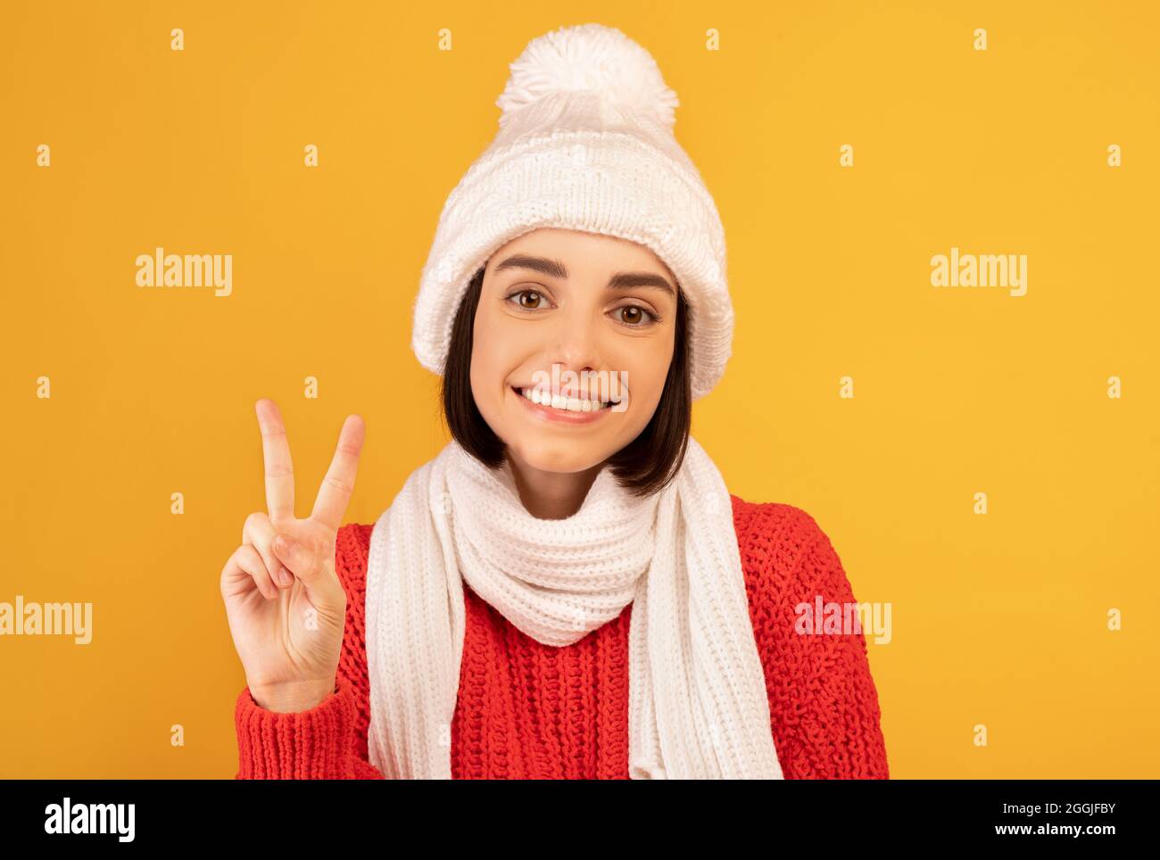 Feliz dama en cálido suéter y sombrero de punto sonriendo y haciendo gesto de paz sobre fondo amarillo del estudio Foto de stock