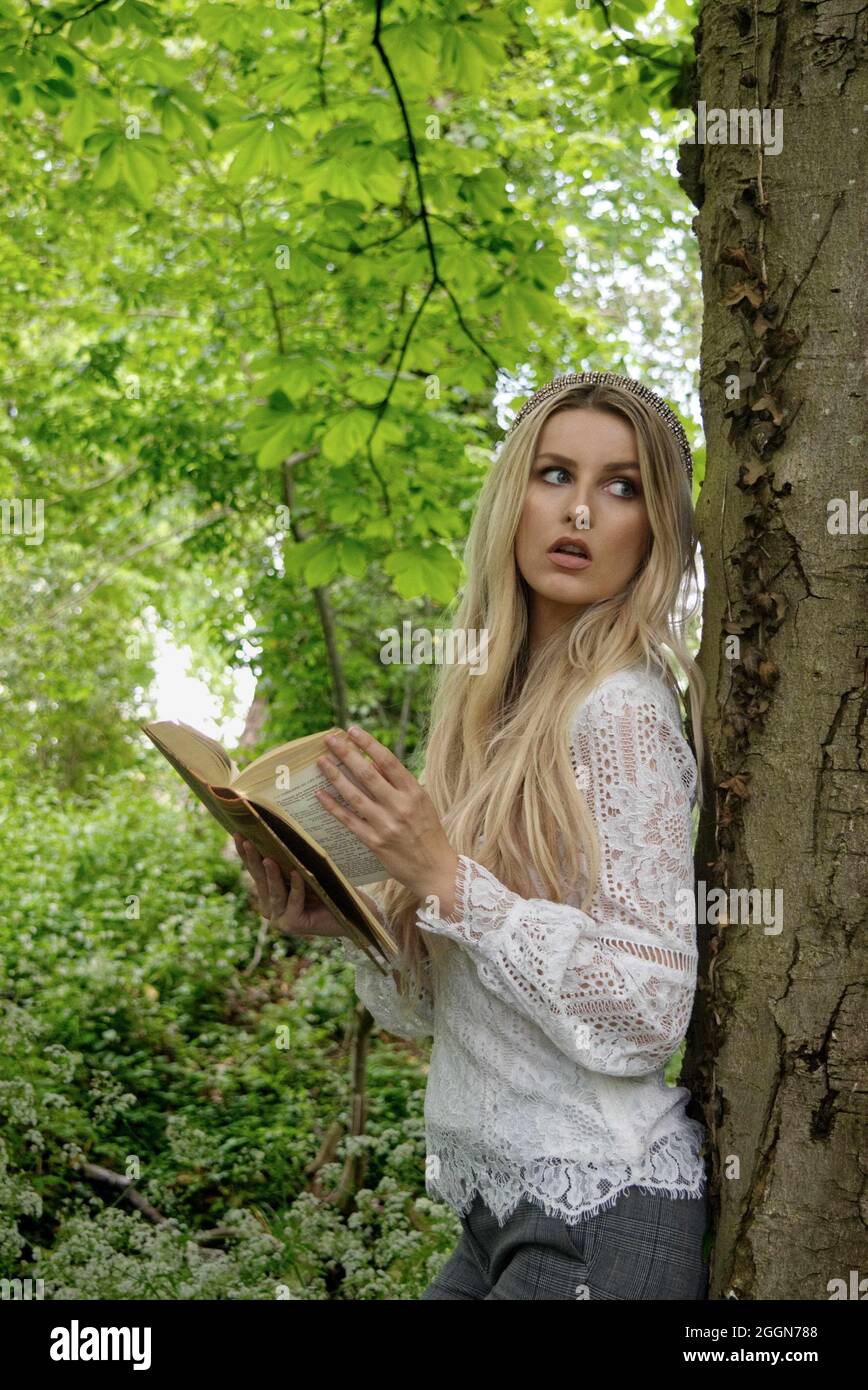 hermosa mujer rubia apoyada contra un árbol sosteniendo un libro, mirando como si perturbada de la lectura Foto de stock