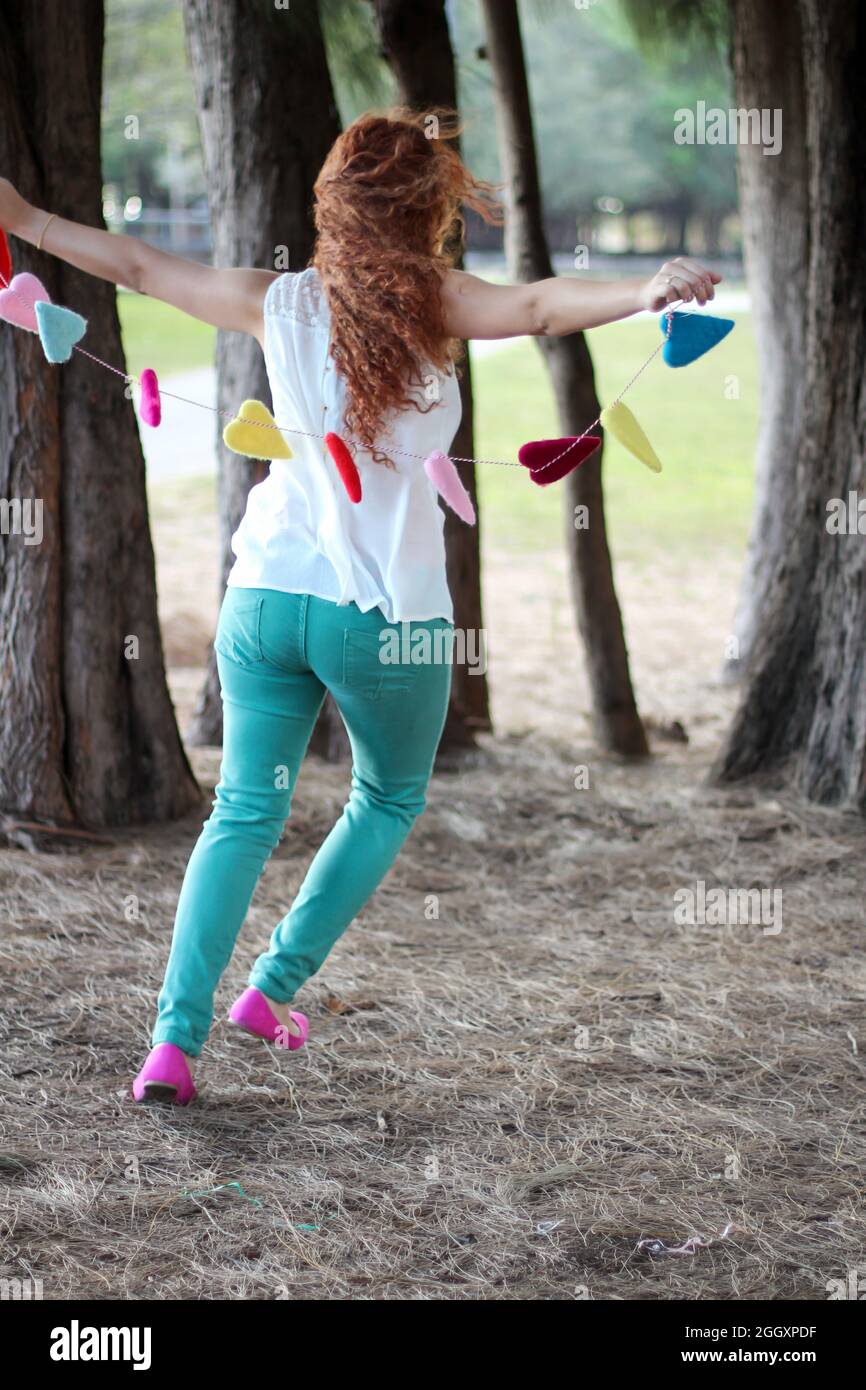 Joven mujer Redhead corriendo en la naturaleza entre árboles que sostienen una guirnalda hecha de corazones para simbolizar amor y romance. Foto de stock