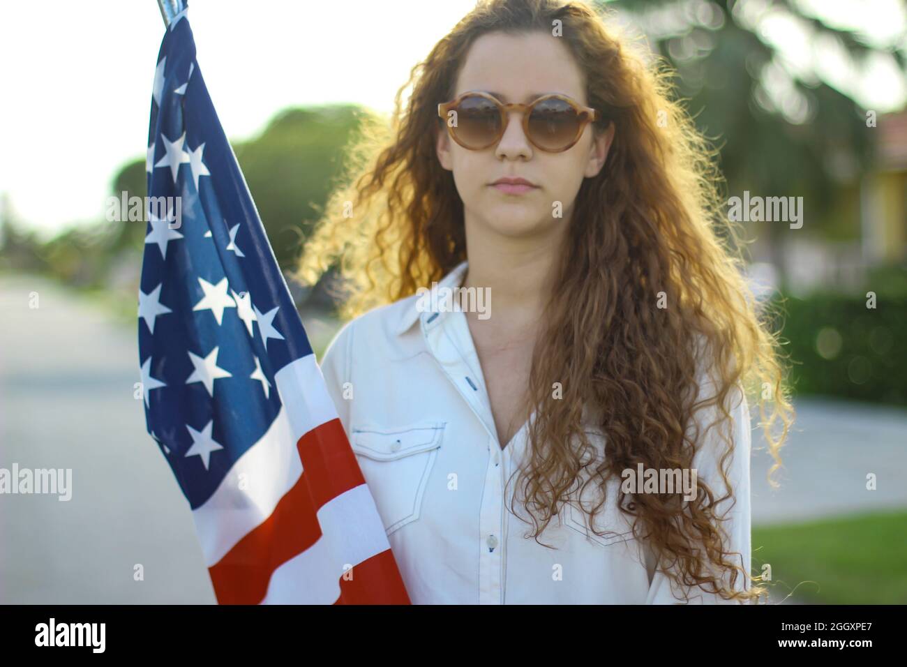 Joven pelirroja hispana y caucásica con gafas de sol sosteniendo la bandera americana afuera. Foto de stock