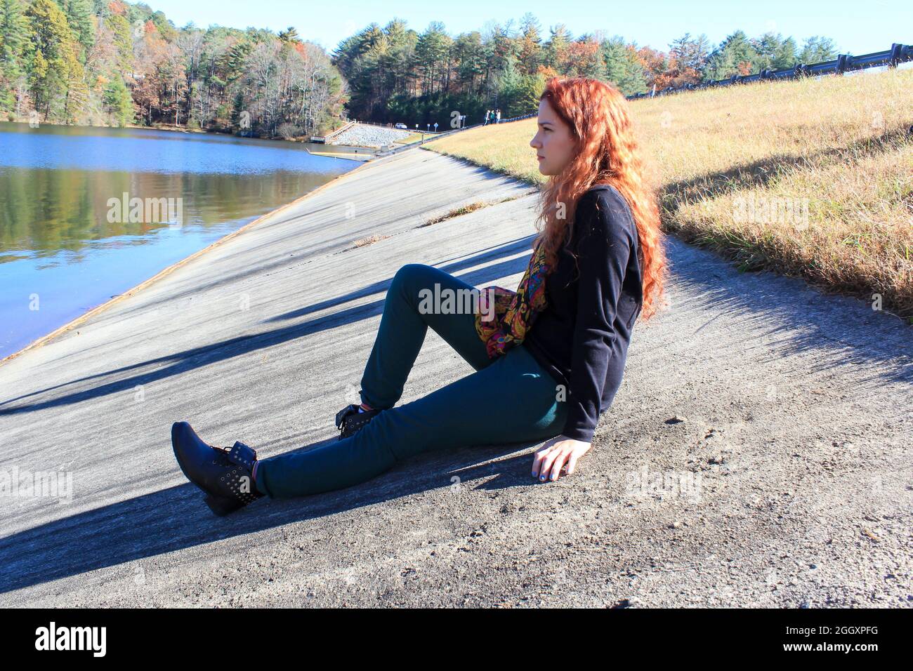 Una mujer joven con pelo rojo está sentada en el suelo junto a un lago en un soleado pero frío día de Georgia. Foto de stock