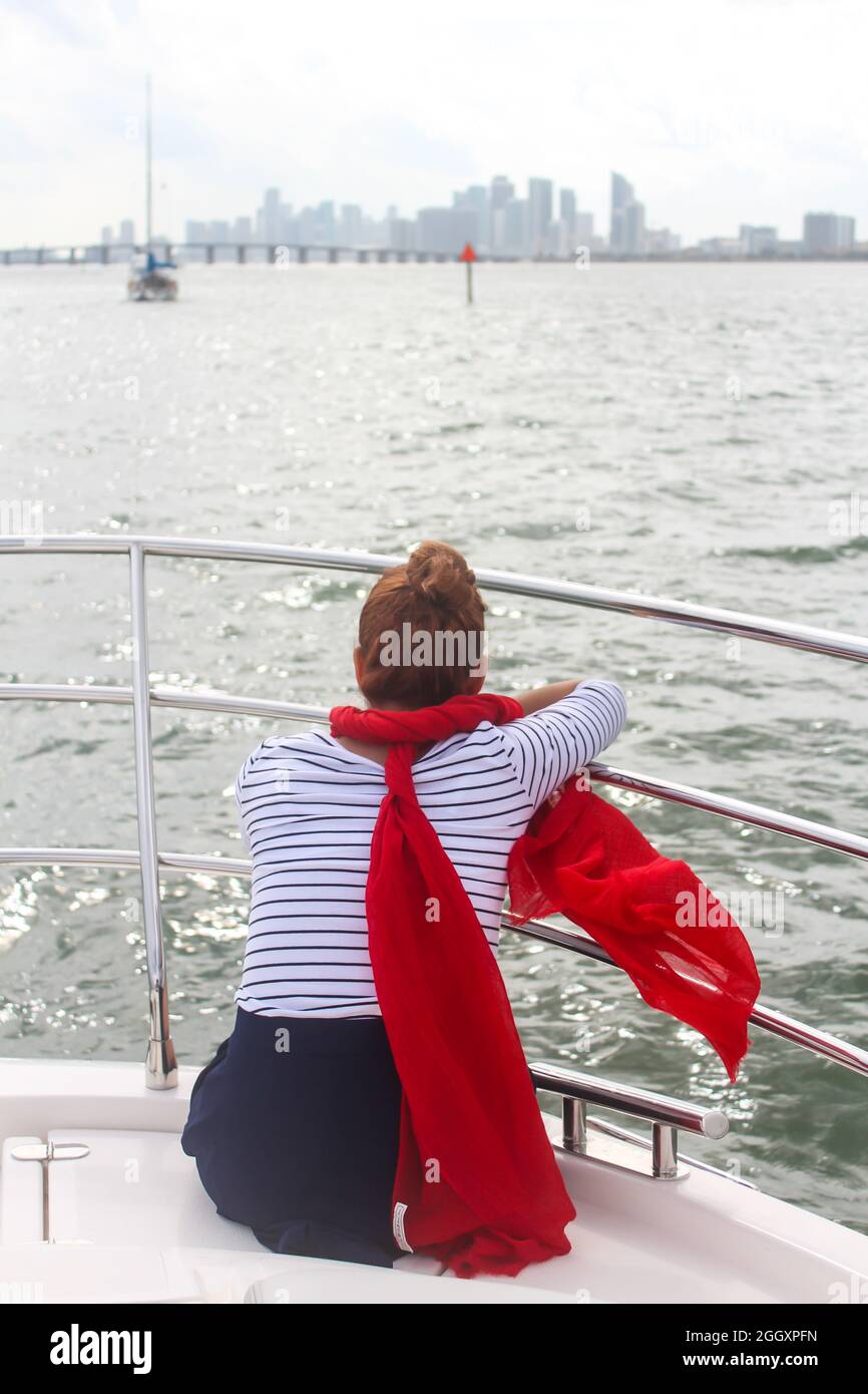 Mujer disfrutando de un lujoso crucero en yate, viaje por el mar en un barco de lujo, mujer con ropa náutica, bufanda roja soplando en el viento y el fondo de la ciudad Foto de stock