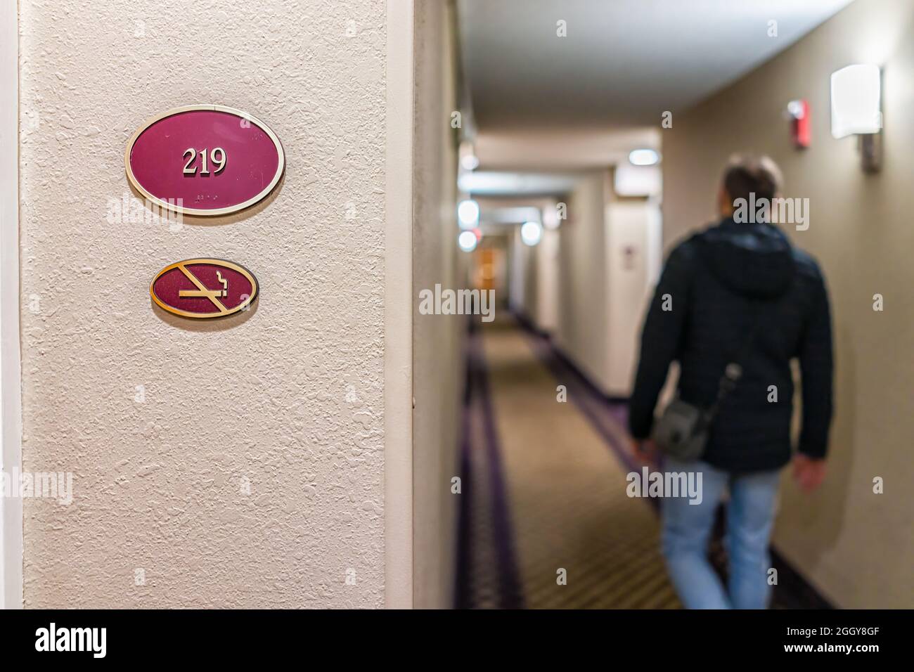 Primer plano de la señal en el hotel motel inn con número de habitación para no fumadores y hombre viajero caminando en el pasillo por la noche Foto de stock