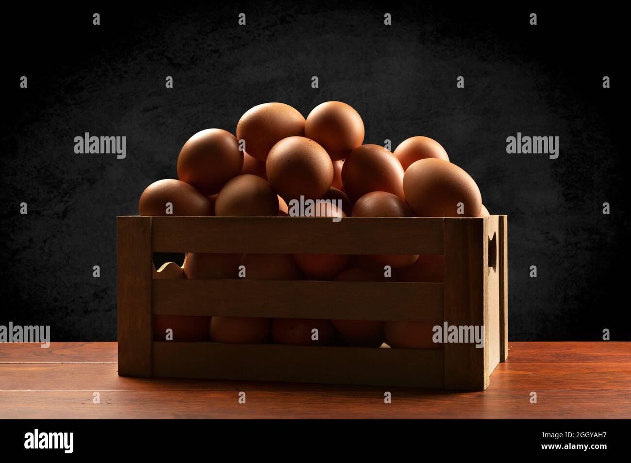 Una pequeña caja marrón de madera de huevos recién recogidos encima de una mesa de teca y enmarcada por un sutil fondo negro moteado. Foto de stock