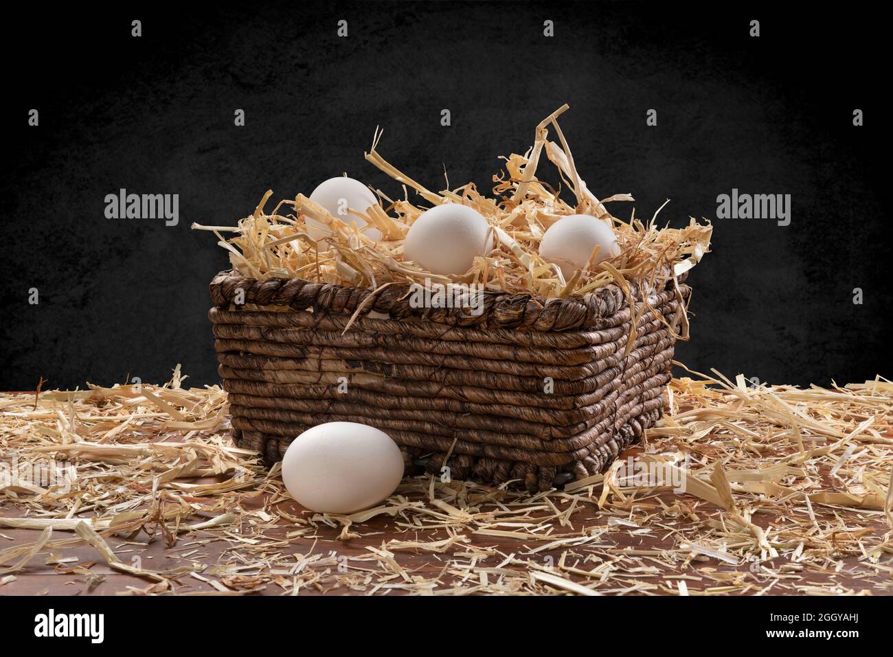 Una pequeña cesta de mimbre de huevos recién recolectados con heno en la parte superior de una mesa de teca y enmarcada por un sutil fondo negro moteado. Foto de stock