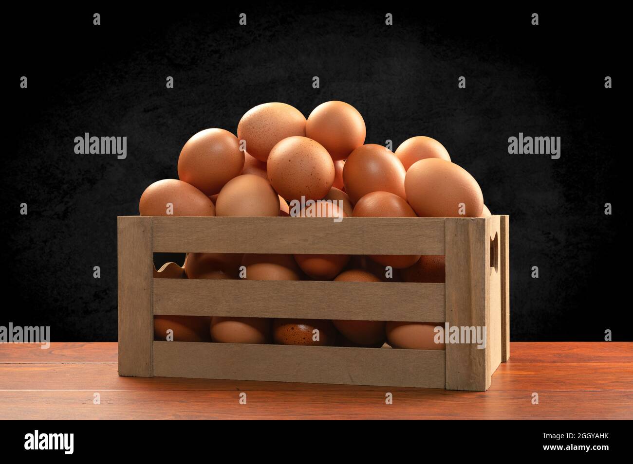 Una pequeña caja marrón de madera de huevos recién recogidos encima de una mesa de teca y enmarcada por un sutil fondo negro moteado. Foto de stock