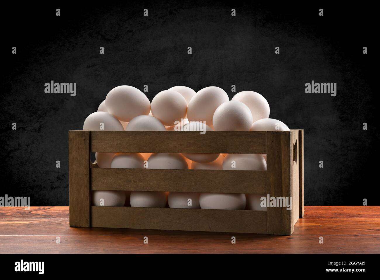 Una pequeña caja marrón de madera de huevos blancos recién recogidos encima de una mesa de teca y enmarcada por un fondo negro sutil, moteado. Foto de stock