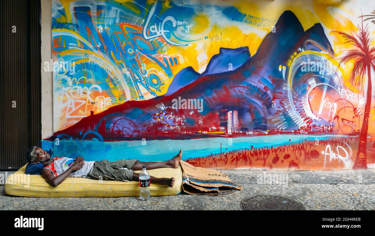 Hombre afrobrasileña duerme rugosa en una calle con un colchón en la parte delantera de un colorido mural que la playa de Ipanema, Río de Janeiro, Brasil Foto de stock