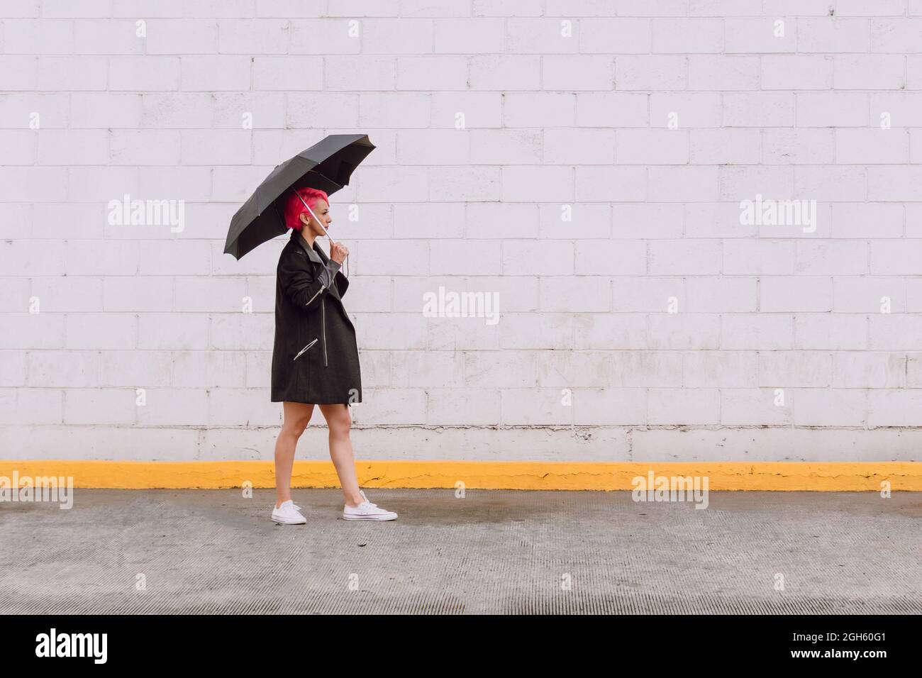 Vista lateral cuerpo completo de mujer joven con pelo rosa en traje de moda  con paraguas y caminar cerca de la pared blanca en la calle Fotografía de  stock - Alamy