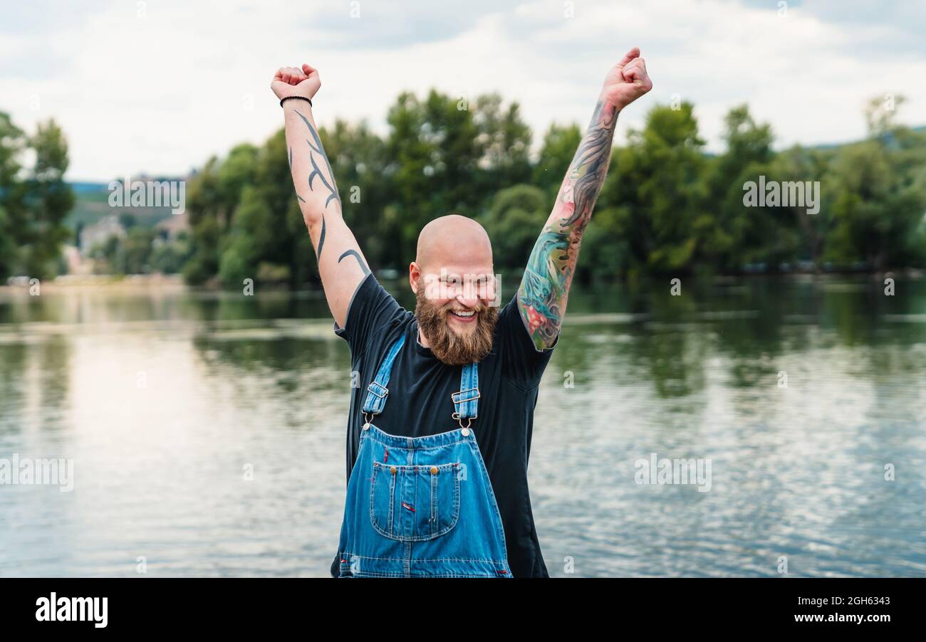 Feliz hombre calvo con barba y tatuajes en ropa informal de pie con brazos elevados cerca del lago y árboles verdes en verano Foto de stock