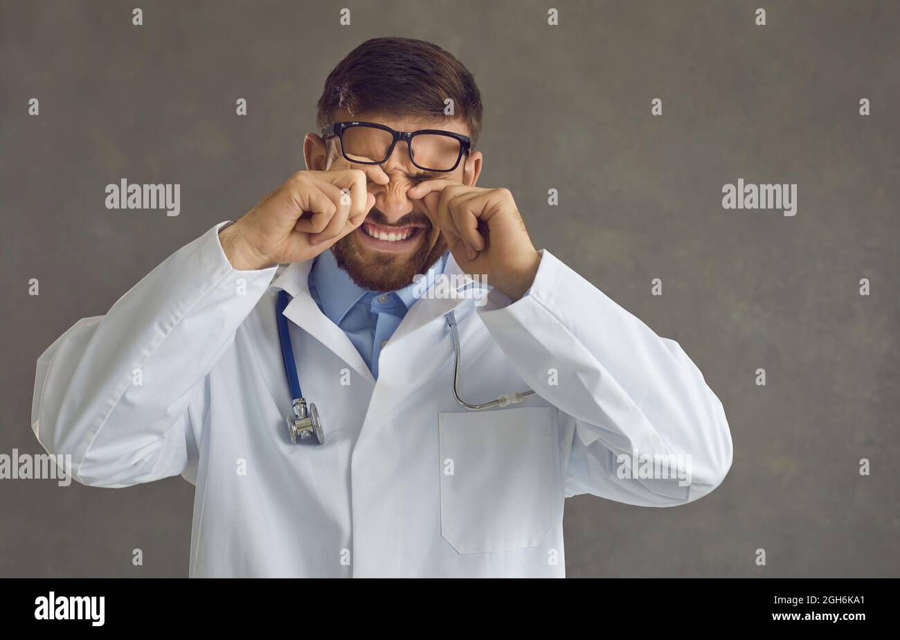 Doctor que está sufriendo fatiga y frotando sus ojos de pie sobre un fondo gris. Foto de stock