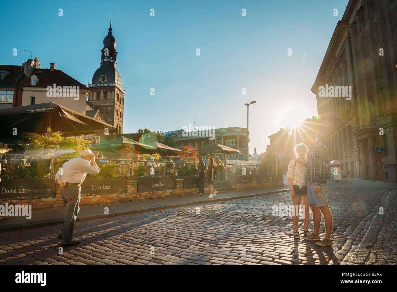 Riga, Letonia. Jóvenes turistas pareja Gente Caminando en la Plaza de la Cúpula en el soleado día de verano. Catedral de la cúpula en el fondo durante la puesta de sol. Foto de stock