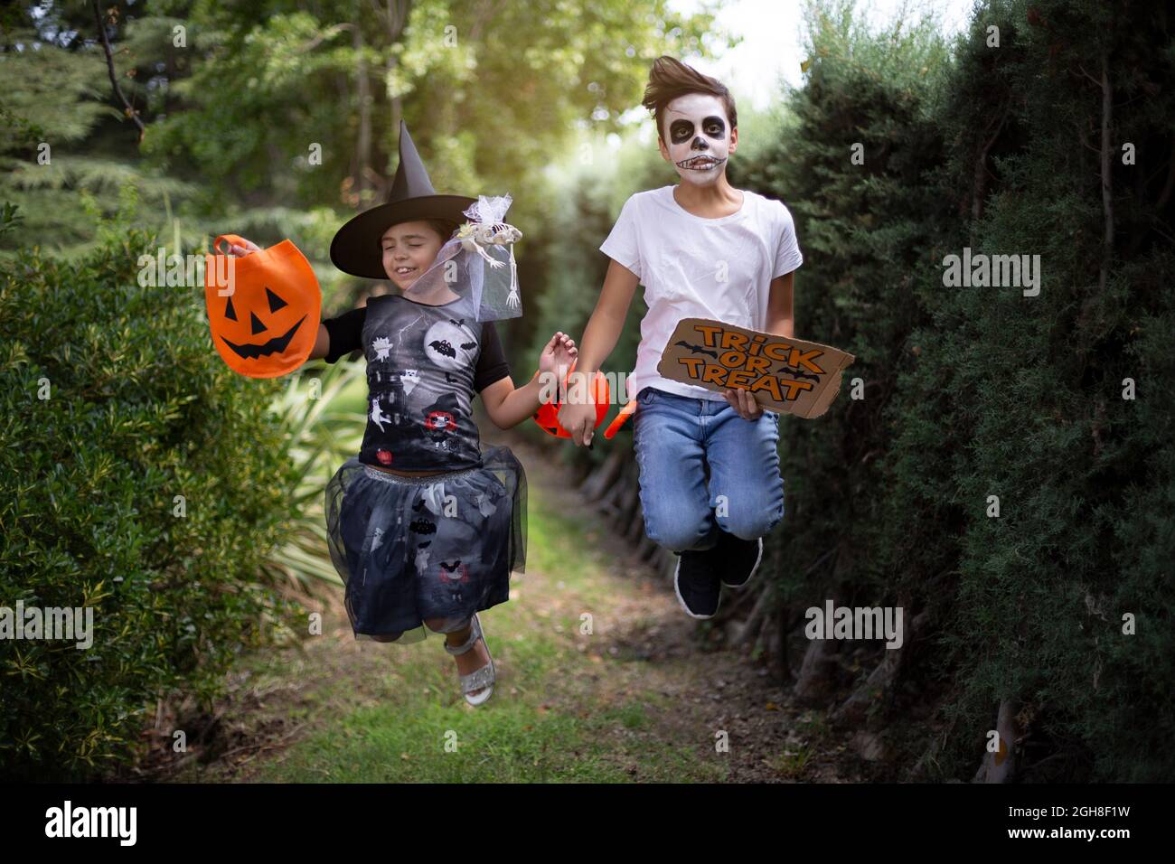 Niño y niña caucásicos con trajes típicos de Halloween saltando y divertirse al aire libre. Foto de stock