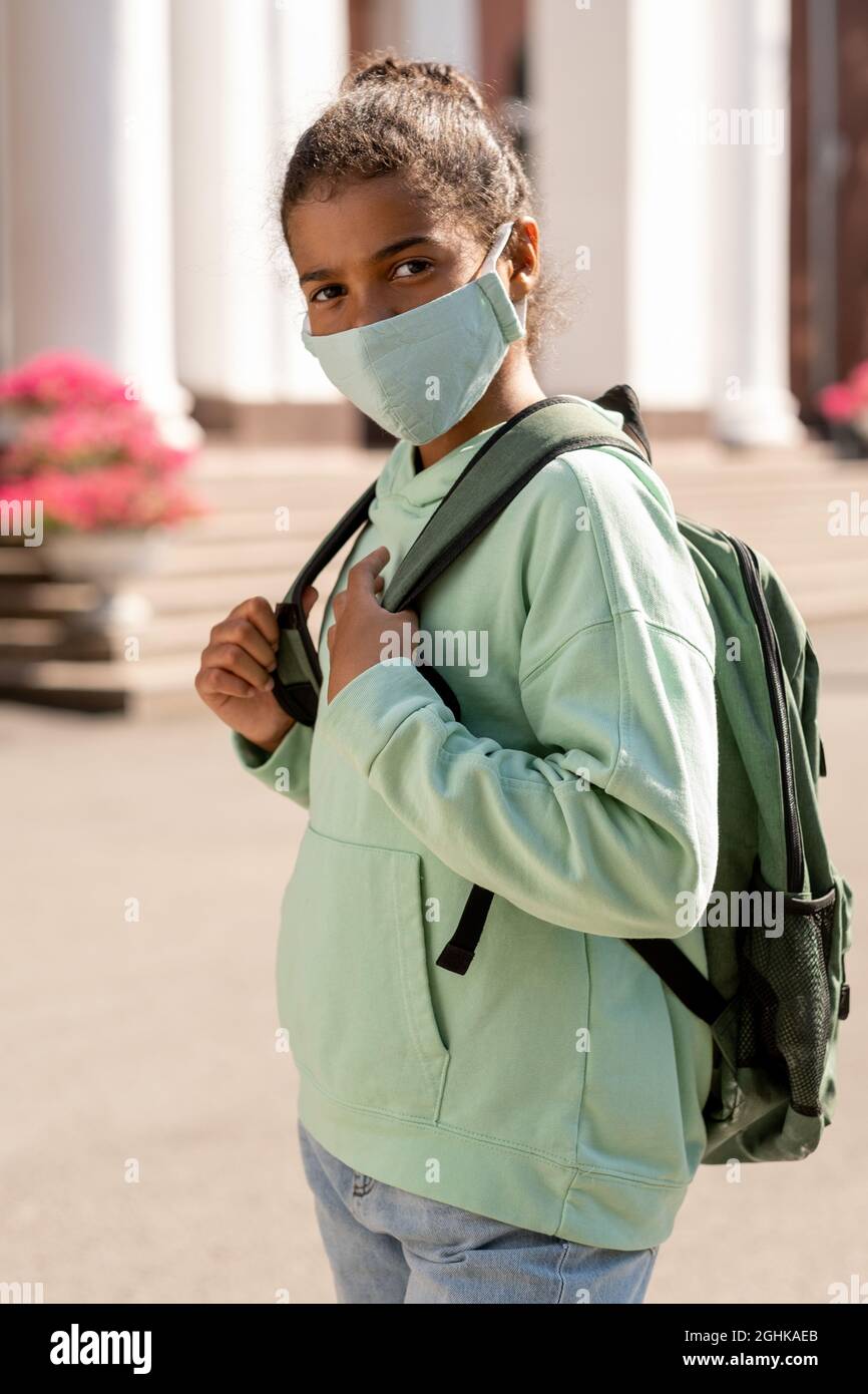 Una linda colegiala afroamericana con mochila con máscara protectora mientras va a la escuela Foto de stock