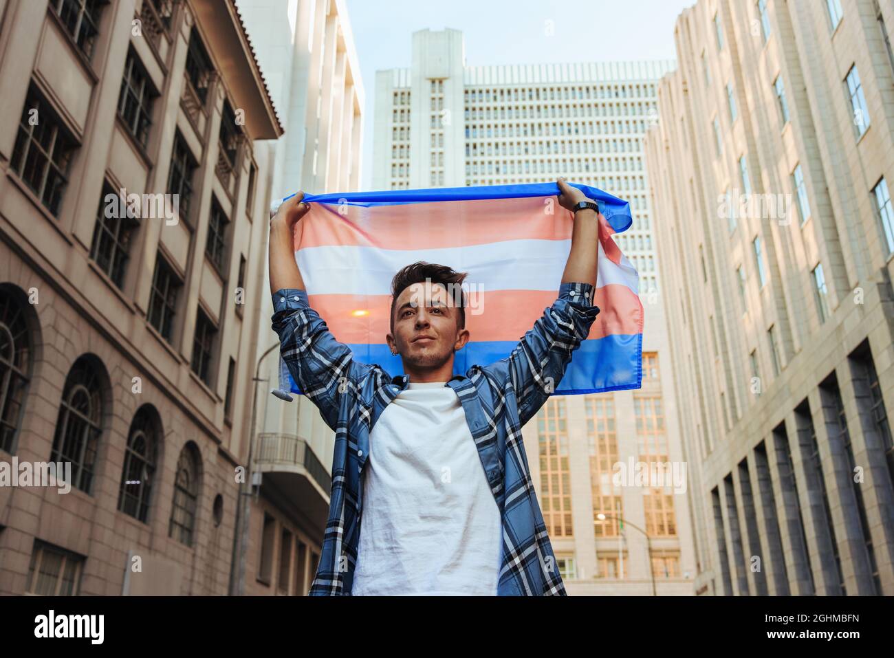 Transman levantando la bandera transgénero al aire libre. Joven transgénero confiado que celebra el orgullo gay en la ciudad. Hombre joven no conforme de género stan Foto de stock