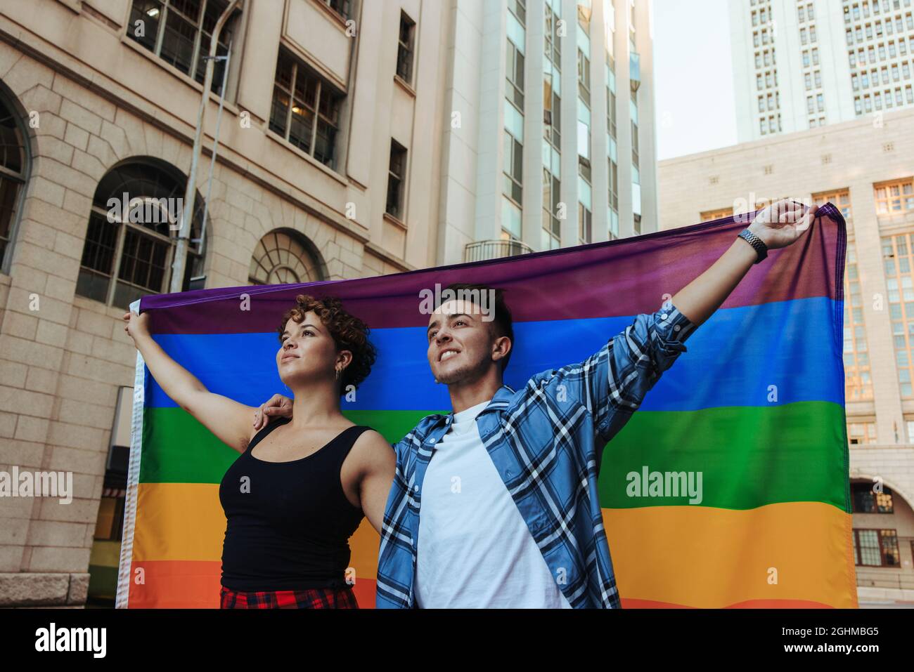Pareja en un desfile de orgullo. Pareja joven y segura de LGBTQ+ levantando la bandera del arco iris mientras se alaban juntos en la ciudad. Pareja joven queer celebrando g Foto de stock