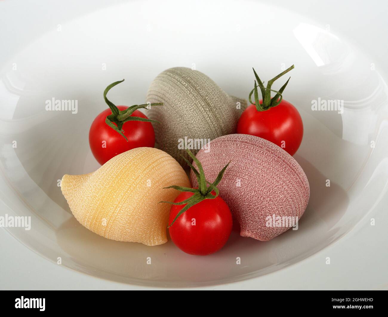 Tres pastas crudas grandes en forma de concha con tres tomates frescos en un plato blanco. Foto de stock