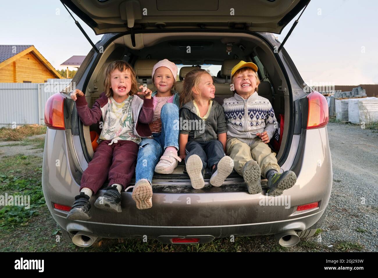 Los niños felices en ropa casual se ríen mientras están sentados en el maletero de automóvil estacionado en la carretera en el campo Foto de stock
