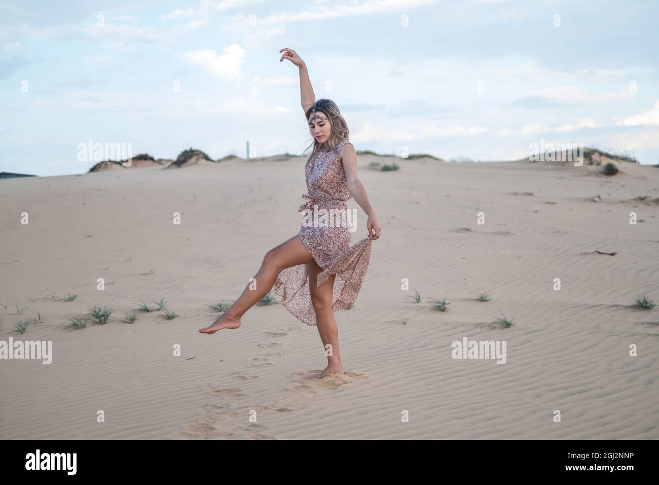 Vista pintoresca de la hermosa mujer rubia en vestido largo casual caminando y bailando en el desierto en día soleado durante las vacaciones de verano. Dunas de arena. Foto de stock