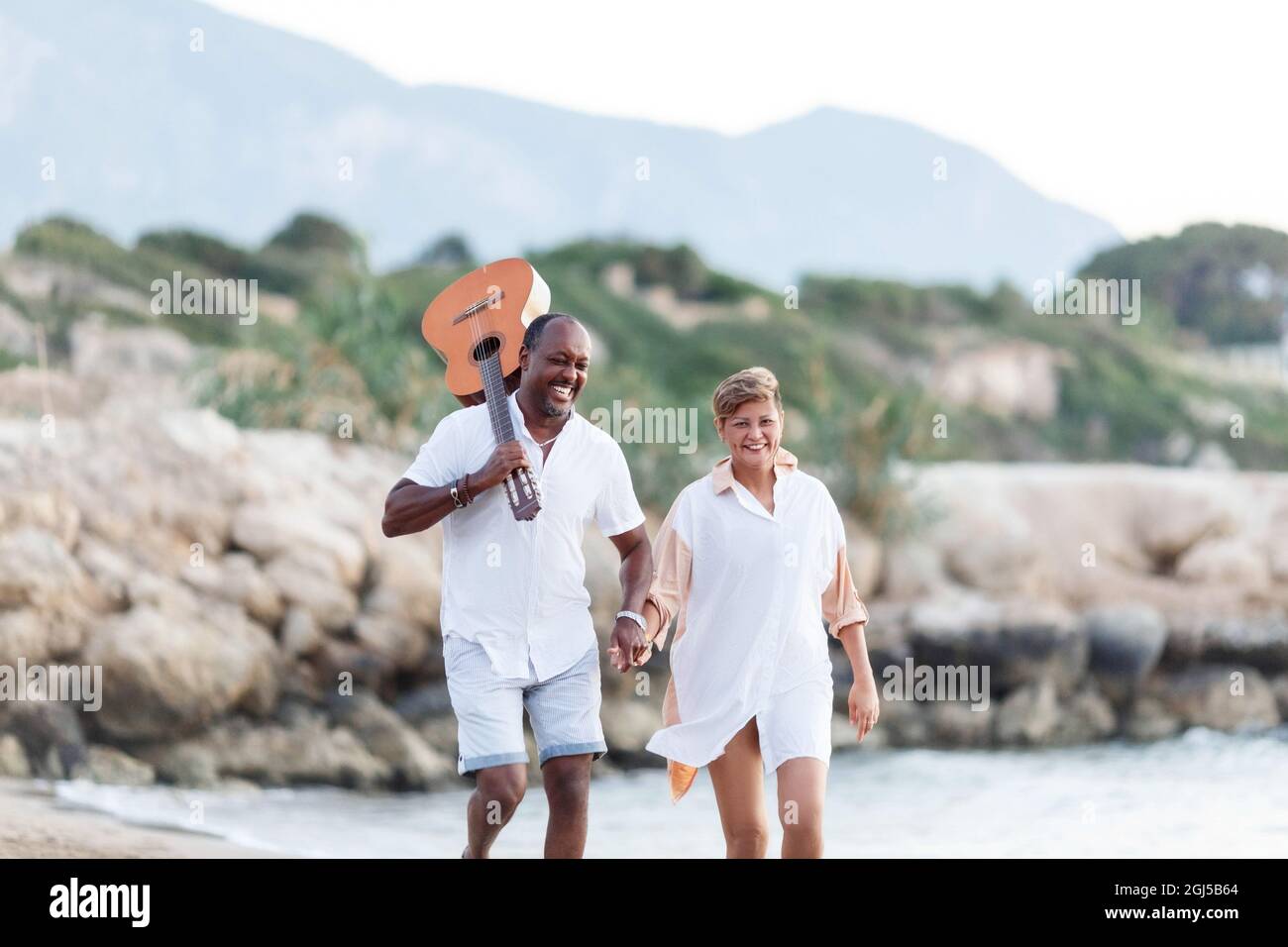 Feliz carrera mixta, pareja de mediana edad abrazando mientras caminan en la playa sosteniendo una guitarra. Hombre sosteniendo la guitarra con mujer. Foto de stock