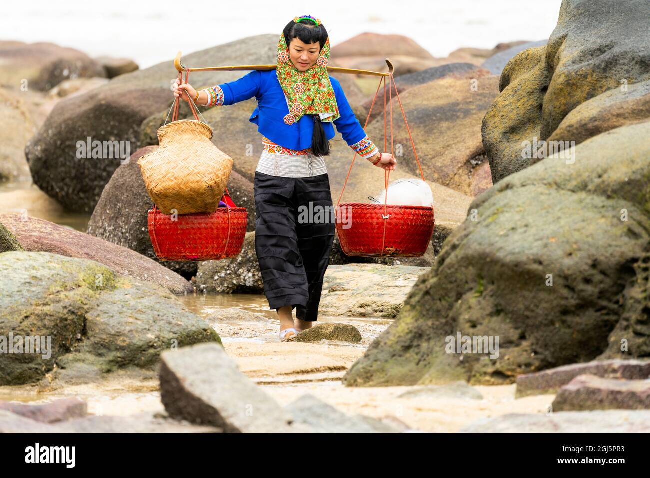 China, Provincia de Fujian, Hui'an. Una mujer con vestido tradicional lleva dos cestas mientras camina sobre las rocas. (Sólo para uso editorial) Foto de stock
