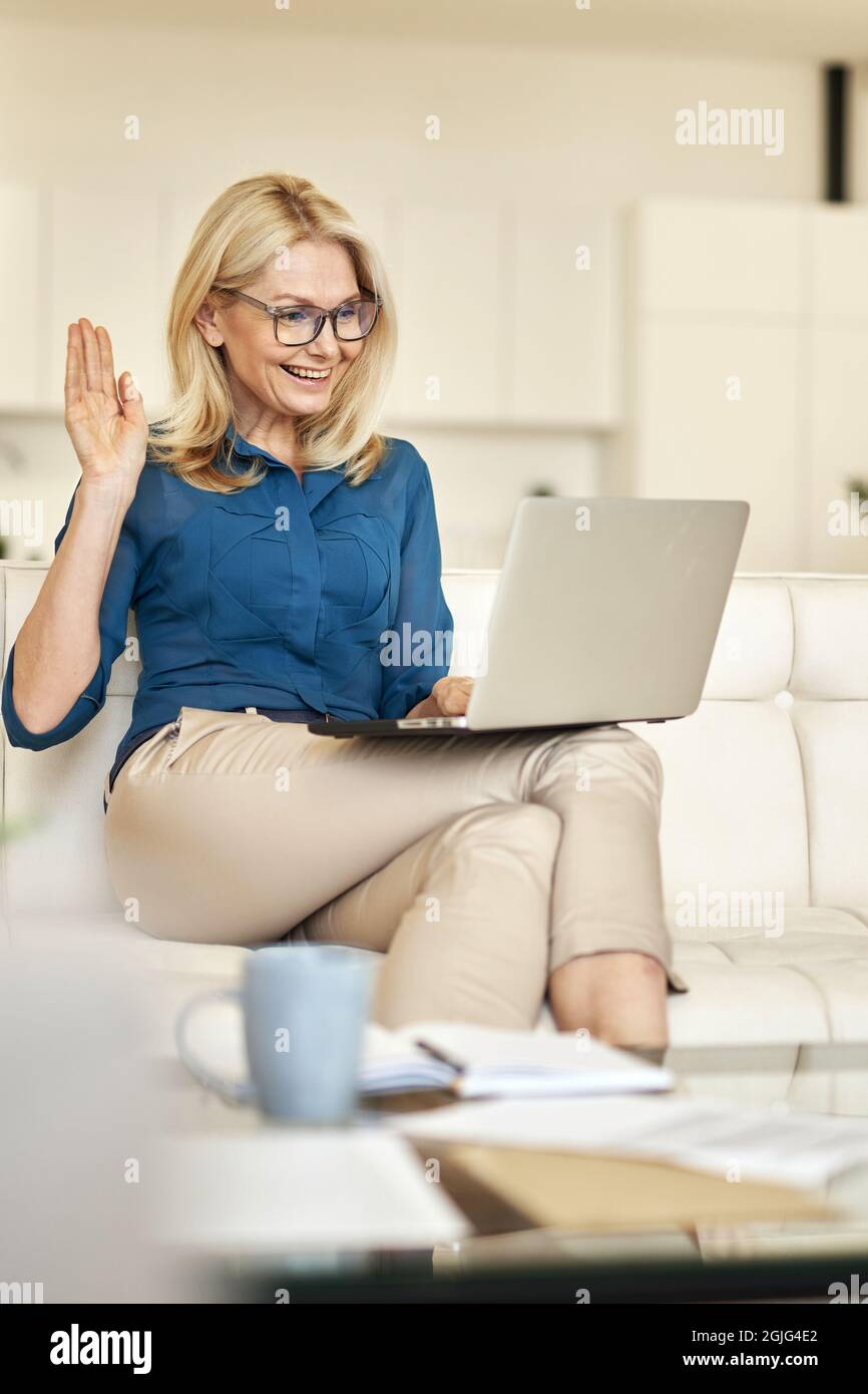 Atractiva mujer madura que trabaja desde casa, agitando la webcam durante  una videollamada en línea con el portátil mientras se sienta en un sofá en  un apartamento moderno Fotografía de stock -