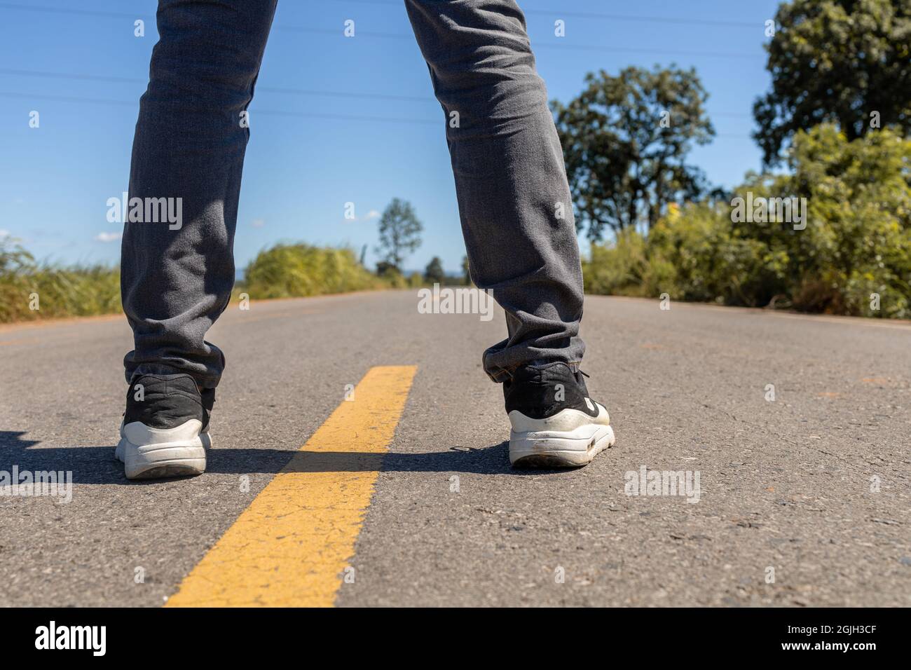 Pies de hombre de pie sobre camino de asfalto con líneas de marcado amarillas. con zapatillas y jeans Fotografía de stock - Alamy