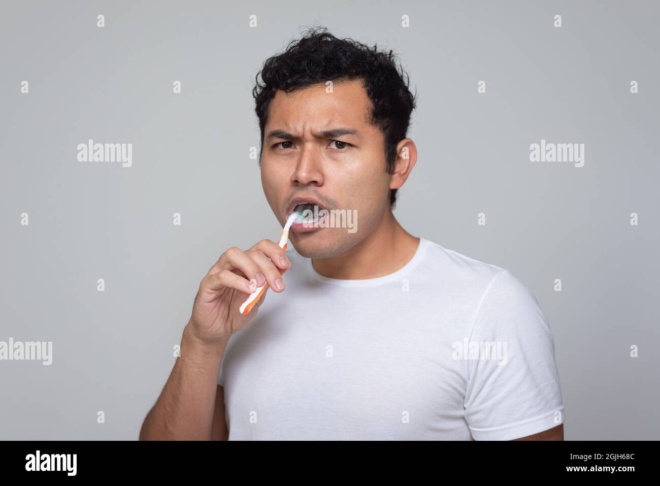 Un hombre mexicano con aspecto latino hispano se cepilla los dientes con un cepillo de dientes naranja, un hombre joven con pelo chino en camisa blanca con fondo gris Foto de stock