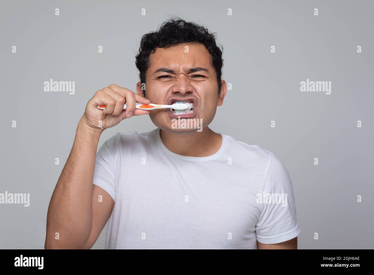 Un hombre mexicano con aspecto latino hispano se cepilla los dientes con un cepillo de dientes naranja, un hombre joven con pelo chino en camisa blanca con fondo gris Foto de stock