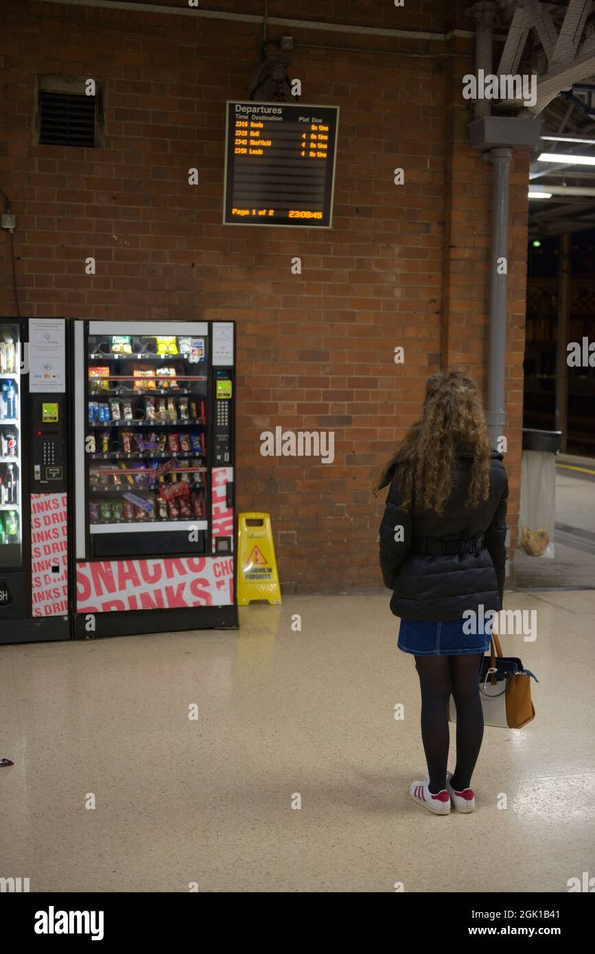 Doncaster, Reino Unido, 22nd de mayo de 2021: Imagen vertical de una joven vestida de invierno leyendo el tablero de salida de la estación de tren de doncaster. Tarde por la noche Foto de stock