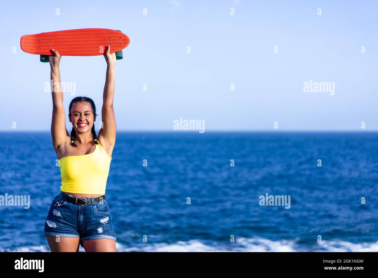 Mujer joven sosteniendo su monopatín sobre un fondo azul del mar. Espacio de copia. Concepto de estilo de vida y deporte Foto de stock