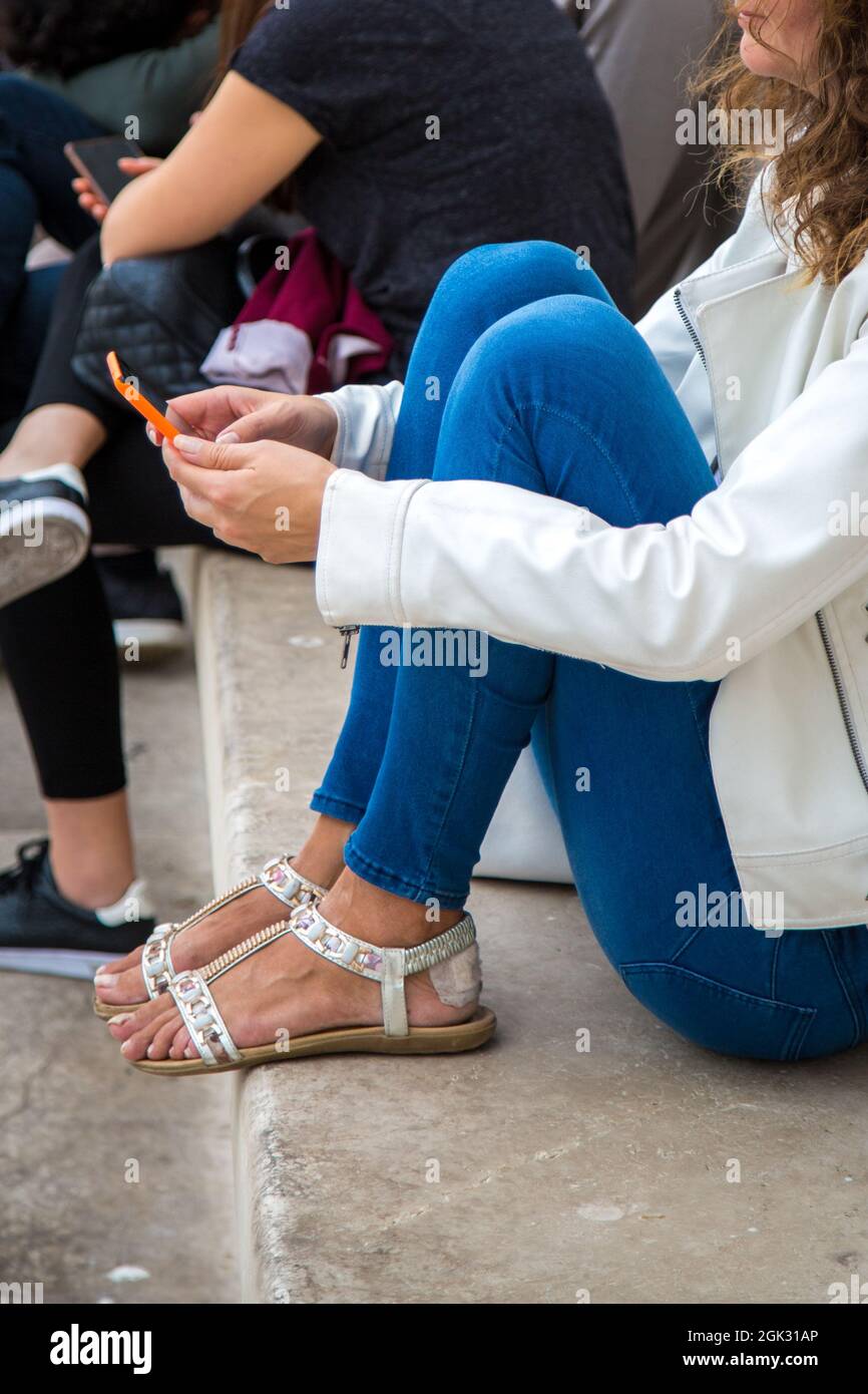 Chica en piel blanca abrigo, azul jeans y sandalias, con yeso en su talón  se sienta y escribe un mensaje en el smartphone al aire libre. Trabajo  inteligente en Fotografía de stock -
