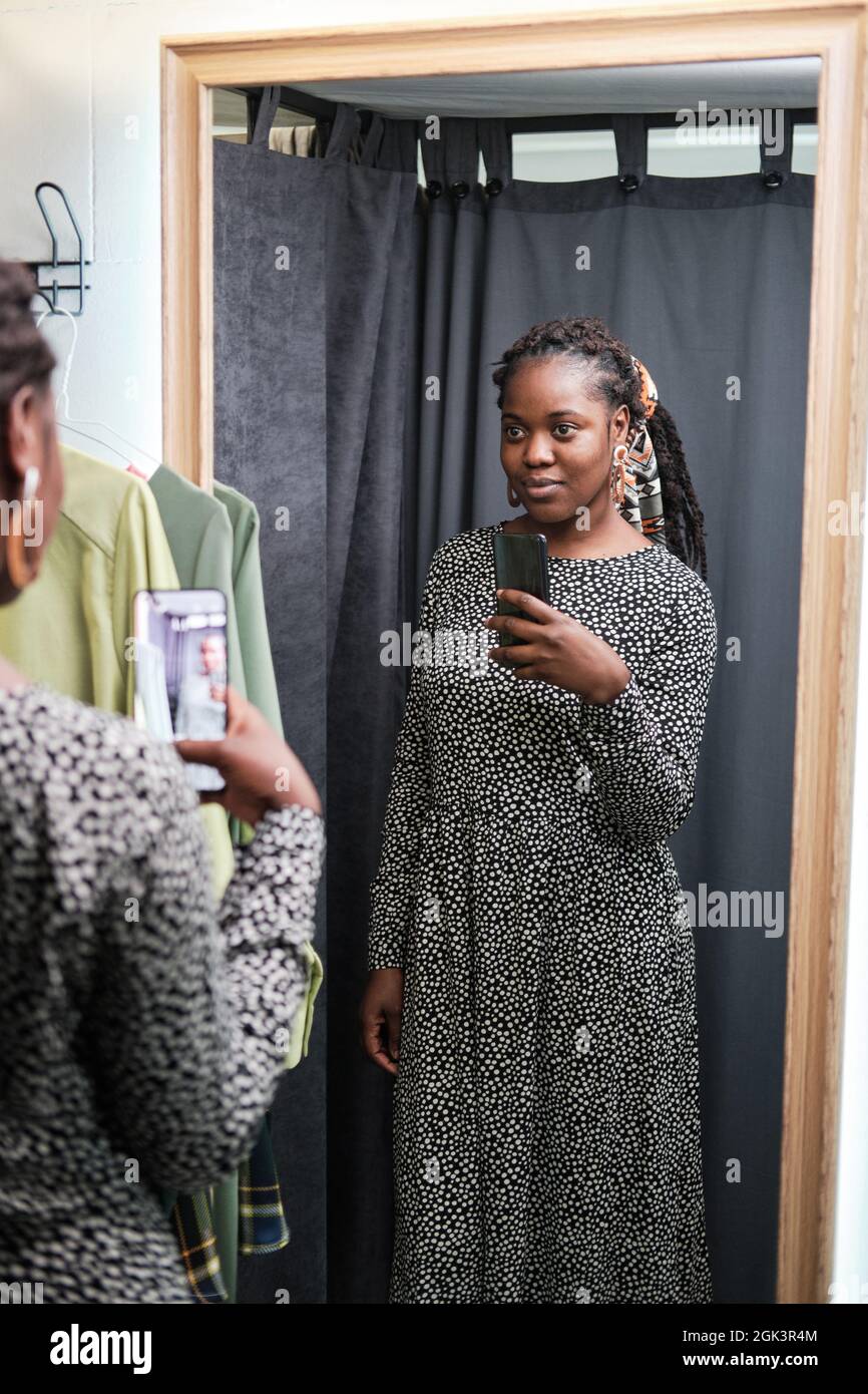Mujer joven africana con nuevo vestido mirando al espejo y haciendo selfie en su teléfono móvil mientras que está de pie en el cuarto de ajuste Foto de stock
