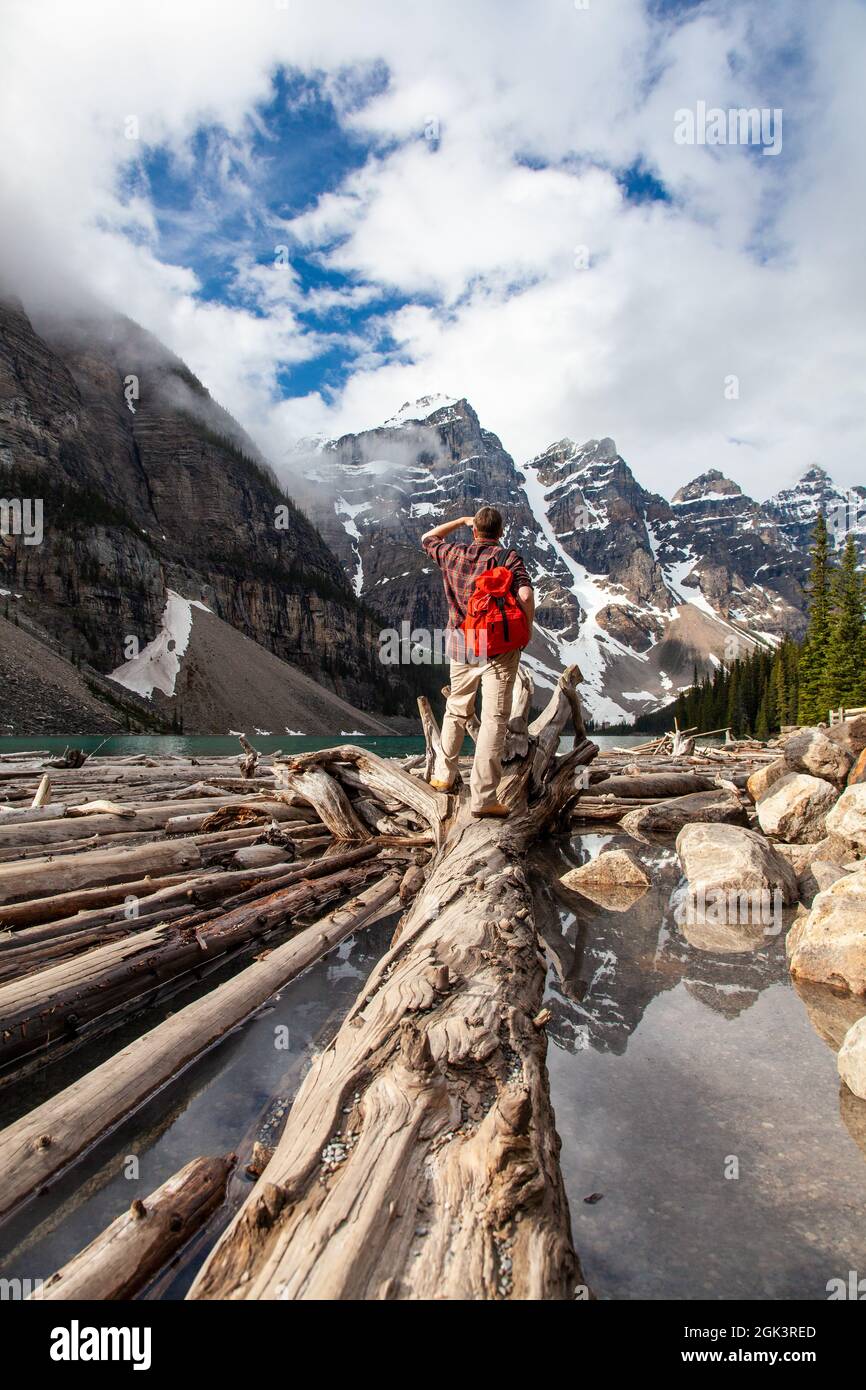 Hombre de senderismo con mochila de rucsac de pie en madera de árbol junto al lago Moraine mirando a los picos cubiertos de nieve de las Montañas Rocosas, Banff National Park, Alberta, Canadá Foto de stock