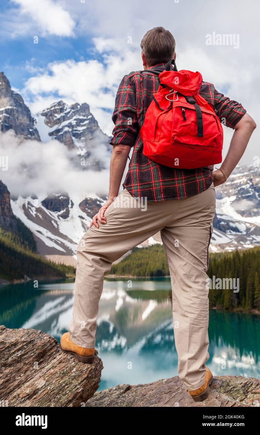Hombre de senderismo con mochila roja en una roca con vistas al lago Moraine mirando a los picos cubiertos de nieve de las Montañas Rocosas, Banff National Park, A. Foto de stock