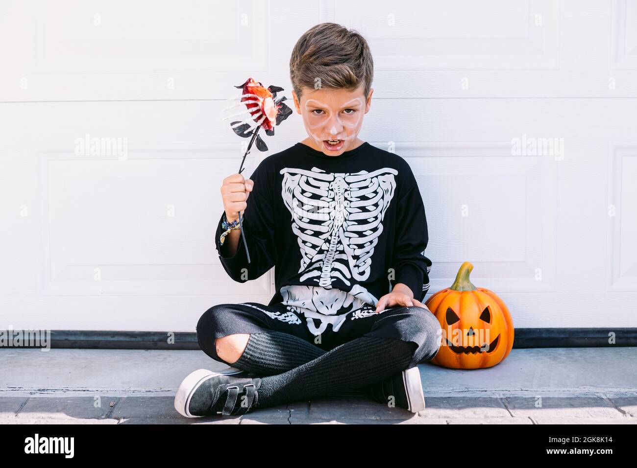 Cuerpo completo de niño preadolescente creepy en traje negro esqueleto con  maquillaje sosteniendo la flor y mirando la cámara mientras se sienta cerca  de la linterna de calabaza contra Fotografía de stock -