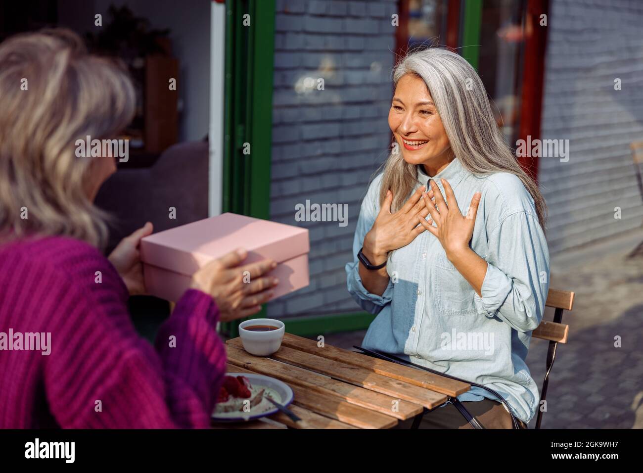 Un amigo maduro da el regalo a una mujer asiática emocional sentada en una pequeña mesa en el café Foto de stock