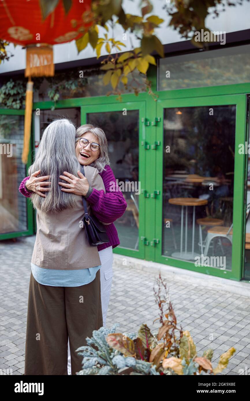 Alegre señora mayor con chaqueta cálida abraza a la reunión de la mejor amiga de pelo largo en la calle de la ciudad Foto de stock