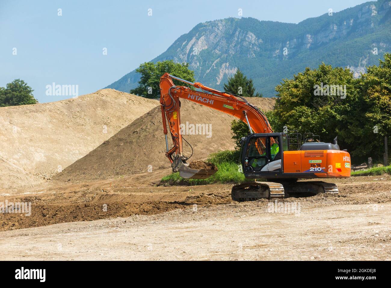 Hitachi digger / maquinaria pesada de planta que trabaja en un sitio, edificio, construcción u otro, en Savoy, Francia. Las montañas se pueden ver en la distancia detrás de este sitio suburbano. (127) Foto de stock