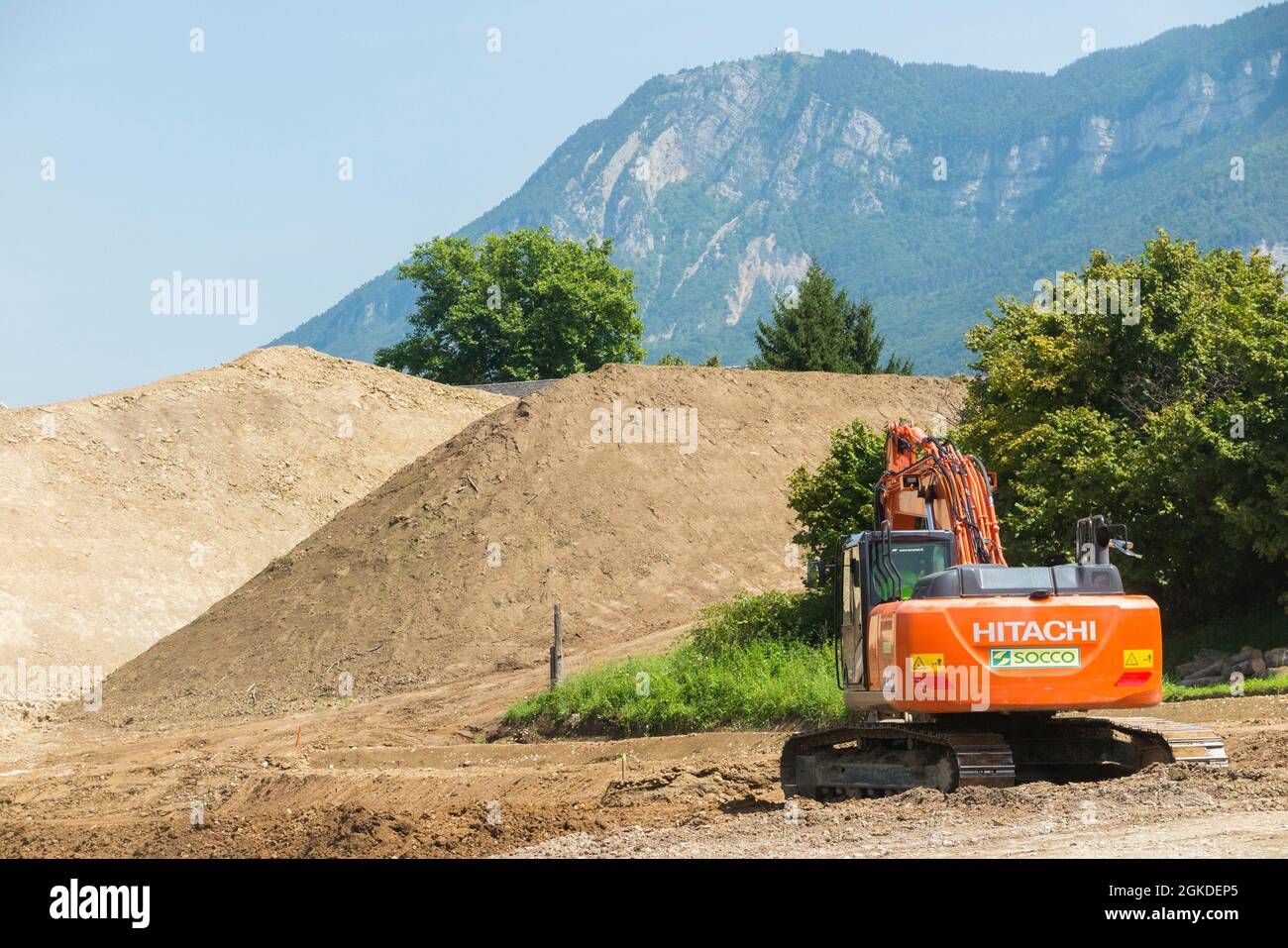 Hitachi digger / maquinaria pesada de planta que trabaja en un sitio, edificio, construcción u otro, en Savoy, Francia. Las montañas se pueden ver en la distancia detrás de este sitio suburbano. (127) Foto de stock