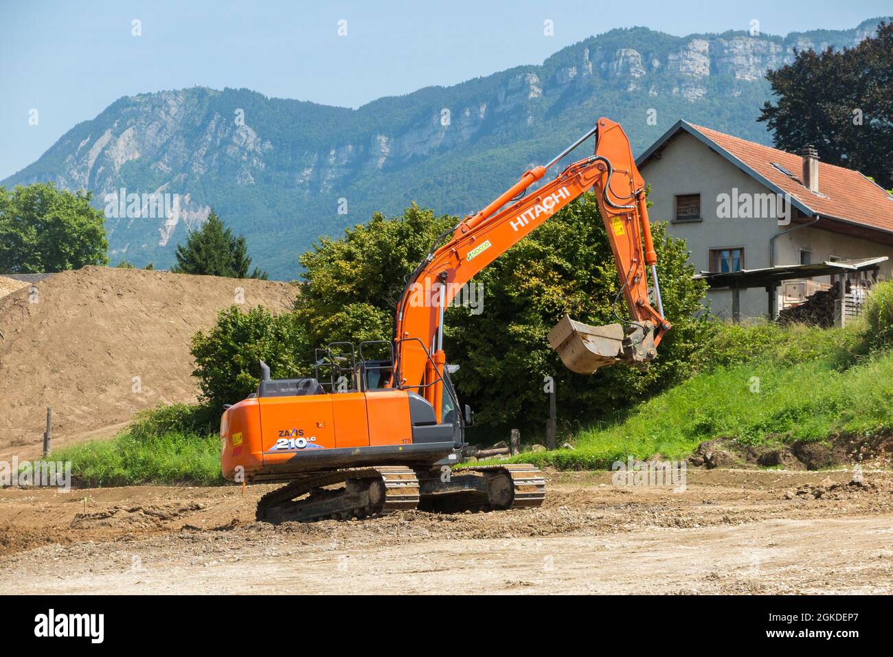 Hitachi digger / maquinaria pesada de planta que trabaja en un sitio, edificio, construcción u otro, en Savoy, Francia. Las montañas se pueden ver en la distancia detrás de este sitio suburbano. (127) Foto de stock