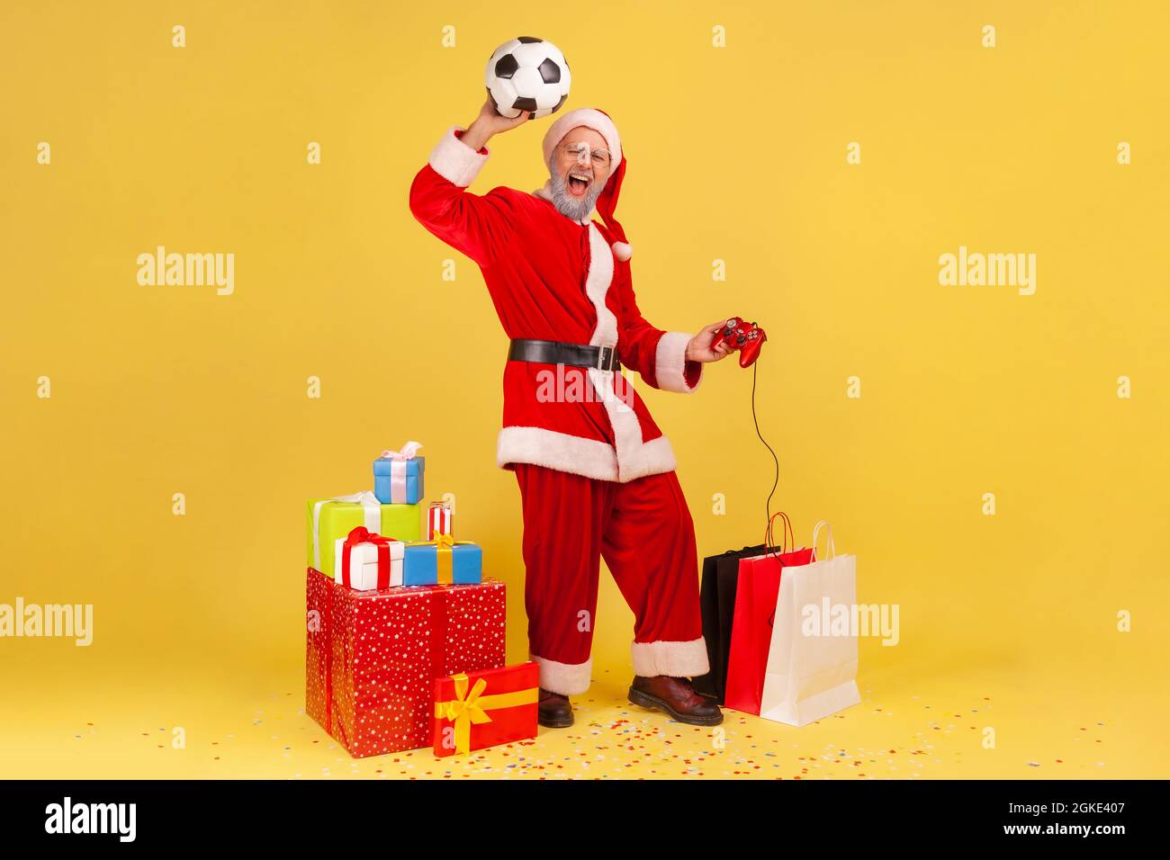 Largo de hombre con barba gris en santa claus traje de brazo elevado con balón de fútbol, con joystick, celebrar ganar en el videojuego de fútbol. YO Foto de stock