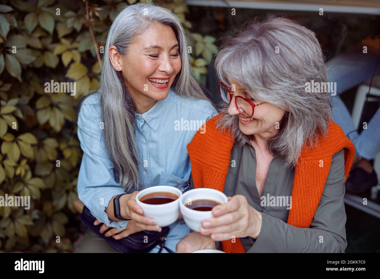 Feliz mujer asiática mayor con tazas de clink de amigo de pelo gris sentado cerca del café al aire libre Foto de stock