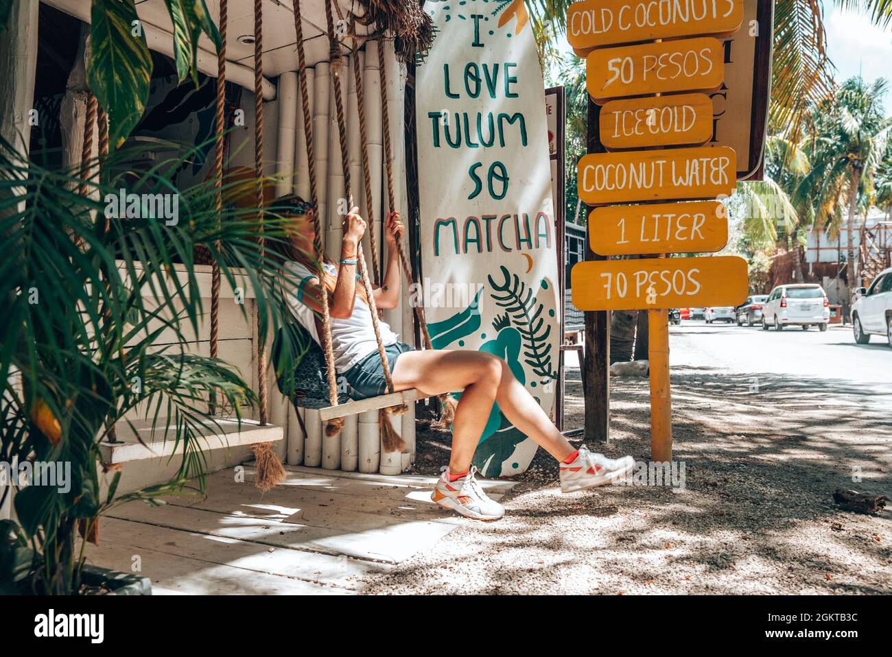 Mujer sentada en el asiento oscilante de la tienda de coco con menú de bebidas al lado de la carretera Foto de stock