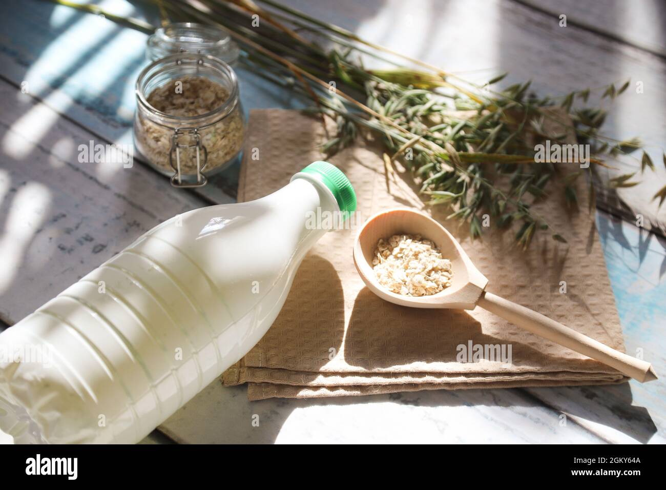 Una cuchara de madera con escamas de avena y una botella de leche de avena sobre la mesa con resplandor solar. Foto de stock