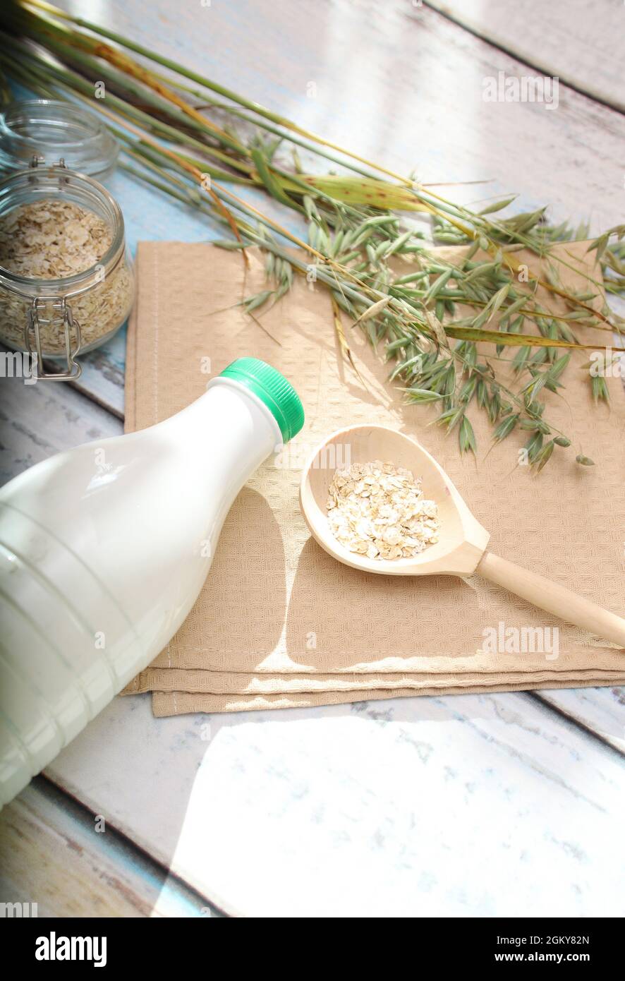 Espigas de avena, una cuchara de madera con escamas de avena y una botella de leche de avena sobre la mesa con resplandor solar. Foto de stock