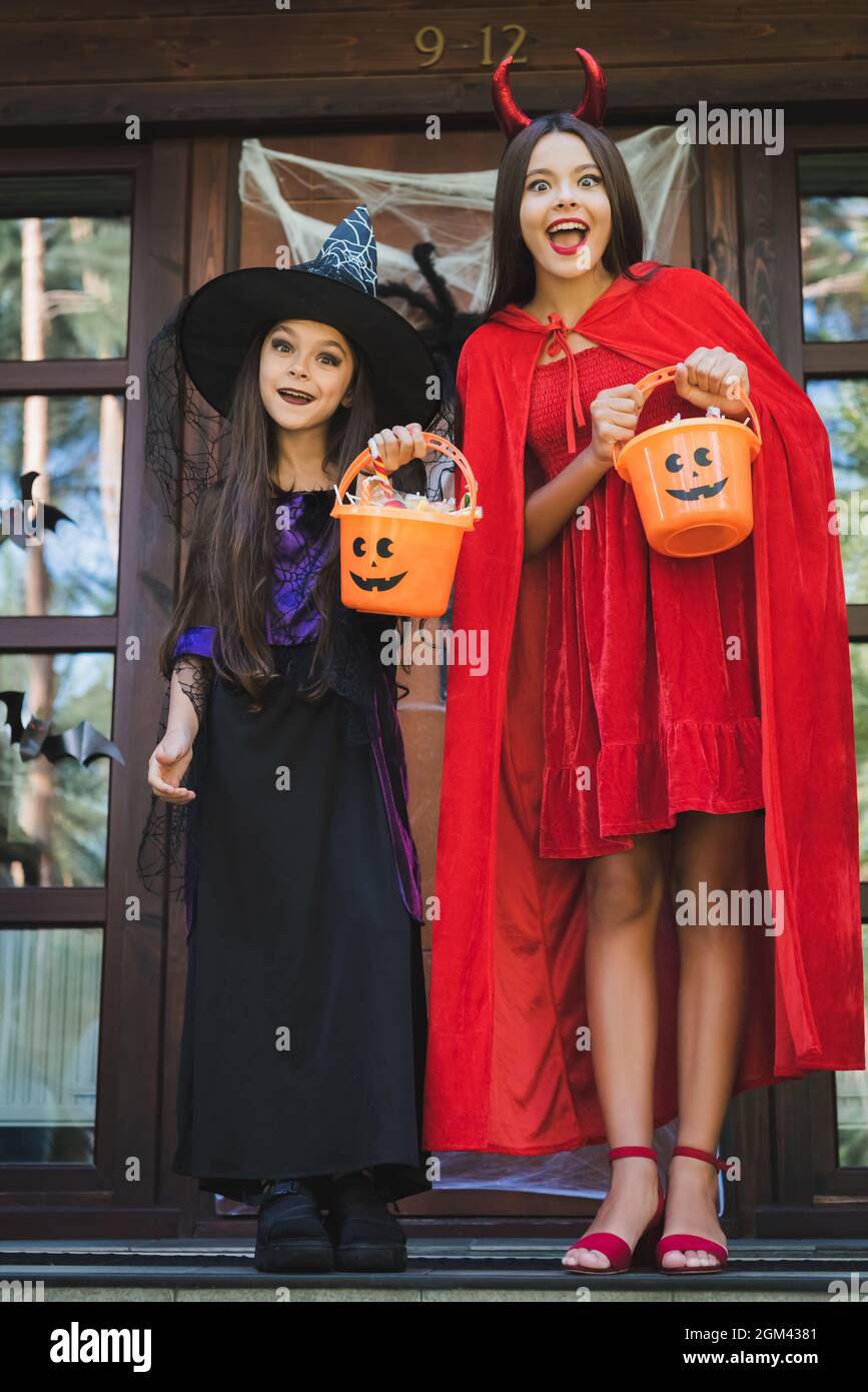 hermanas asombradas en trajes de halloween bruja y diablo, con cubos de  dulces en el porche de la cabaña Fotografía de stock - Alamy