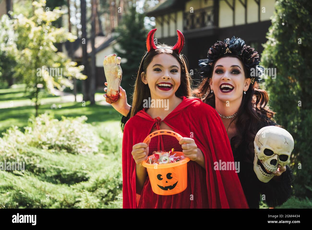 emocionado mamá e hija en disfraces de halloween diablo y vampiro, con  cráneo, mano de juguete y cubo de caramelos Fotografía de stock - Alamy