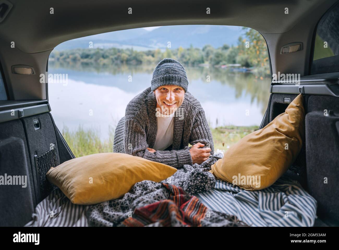 Un hombre de mediana edad con una sonrisa sincera vestido con ropa de punto cálido y jeans, retrato en el acogedor maletero del coche y con hermosas vistas al lago de montaña. Cálido Foto de stock